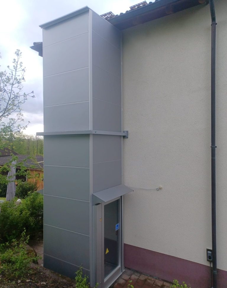 Außenansicht eines modernen Aufzugs im Gebäude, daneben Pflanzen und ein gepflegter Garten.